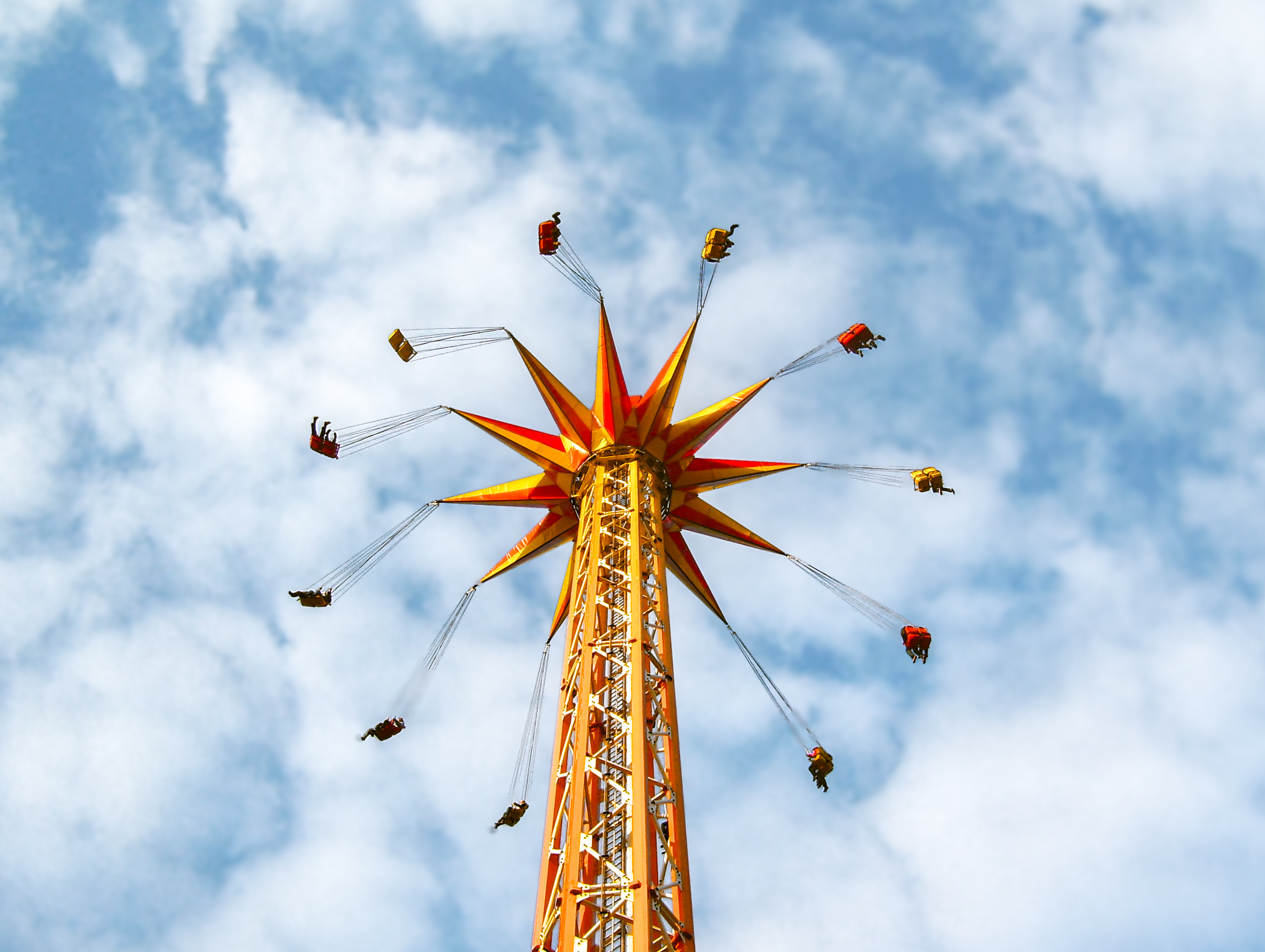 Vibrant image of a tall chair swing amusement ride, captured from below against a partly cloudy blue sky. Colorful chairs with silhouetted riders in motion, evoking feelings of summer, joy, and advent